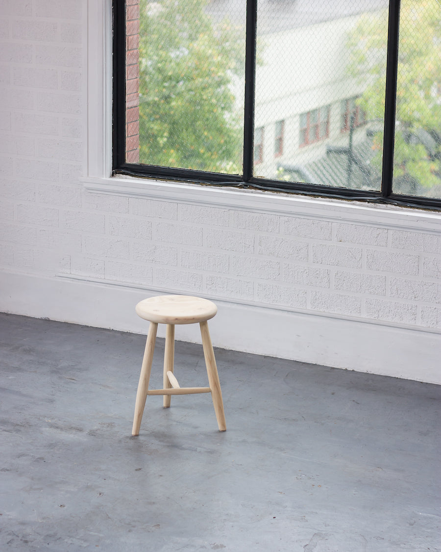 Wooden stool in a room with large windows showing greenery outside.