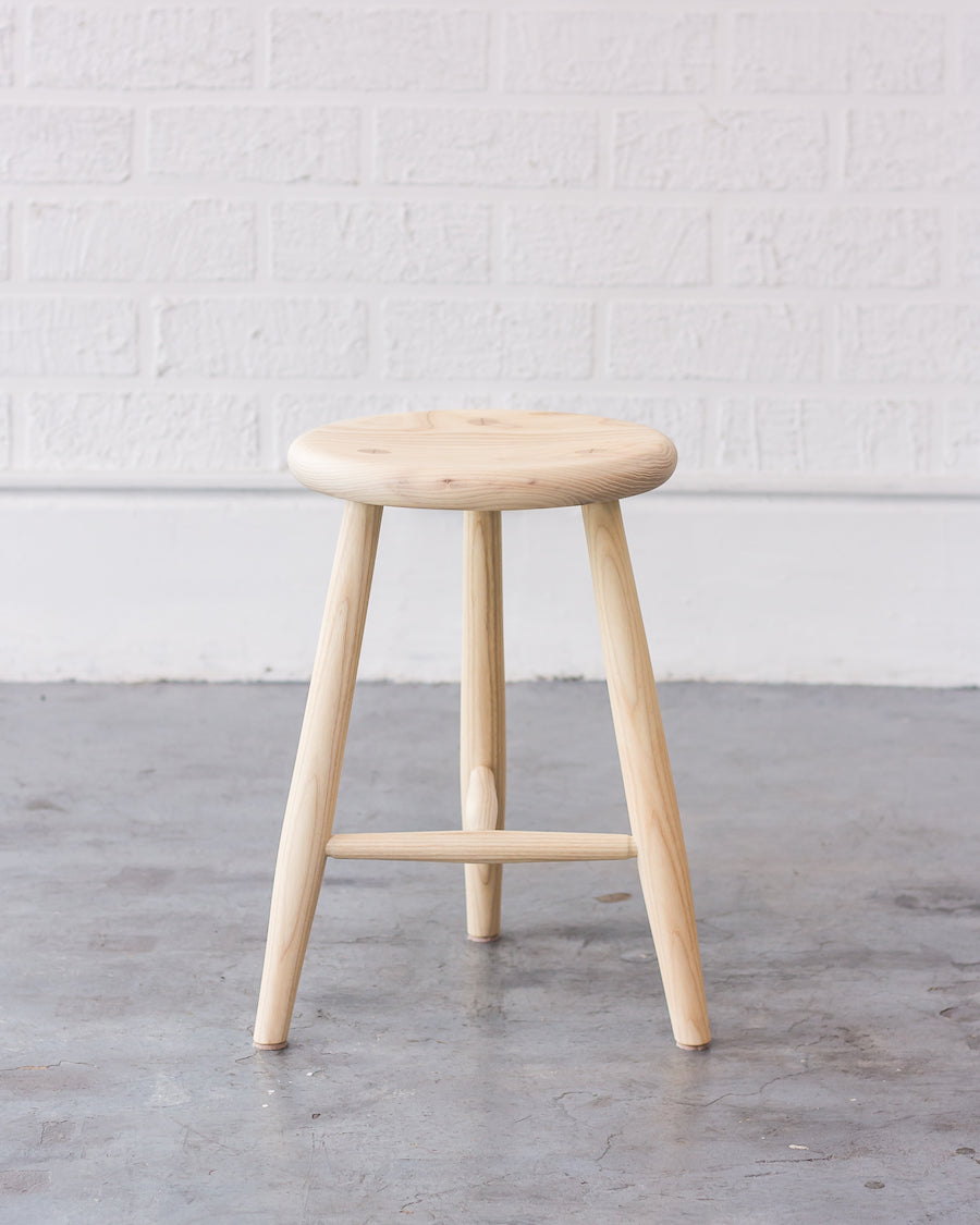 Wooden stool with a round seat and four legs on a gray floor against a white brick wall.