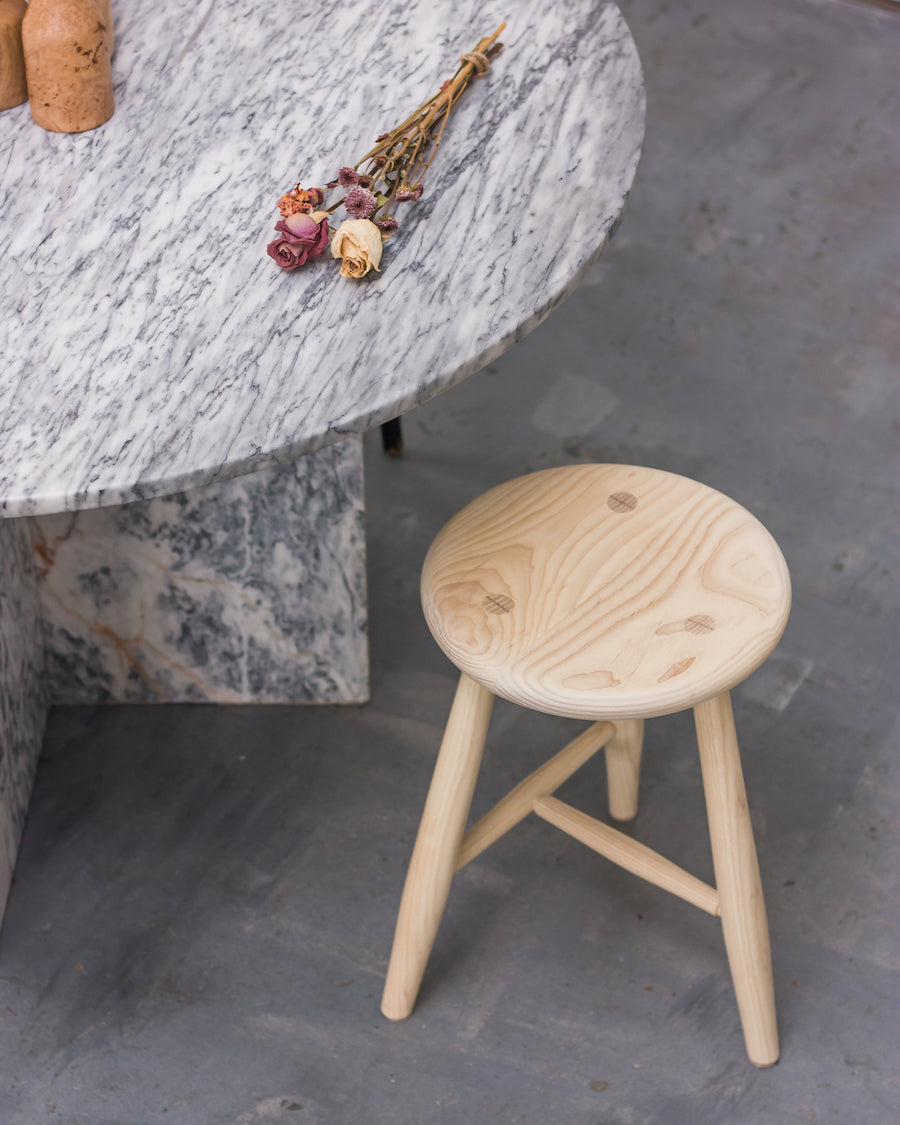 Wooden stool in front of a marble table with dried flowers on a concrete floor.