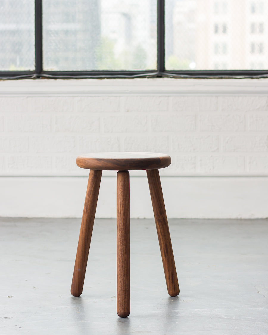 Wooden stool with three legs in a minimalistic room with large windows.