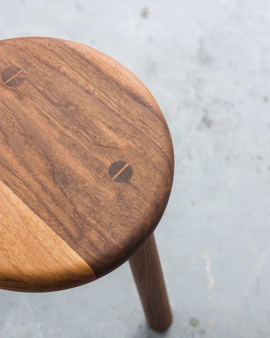 Wooden stool with a light and dark wood finish on a concrete floor.