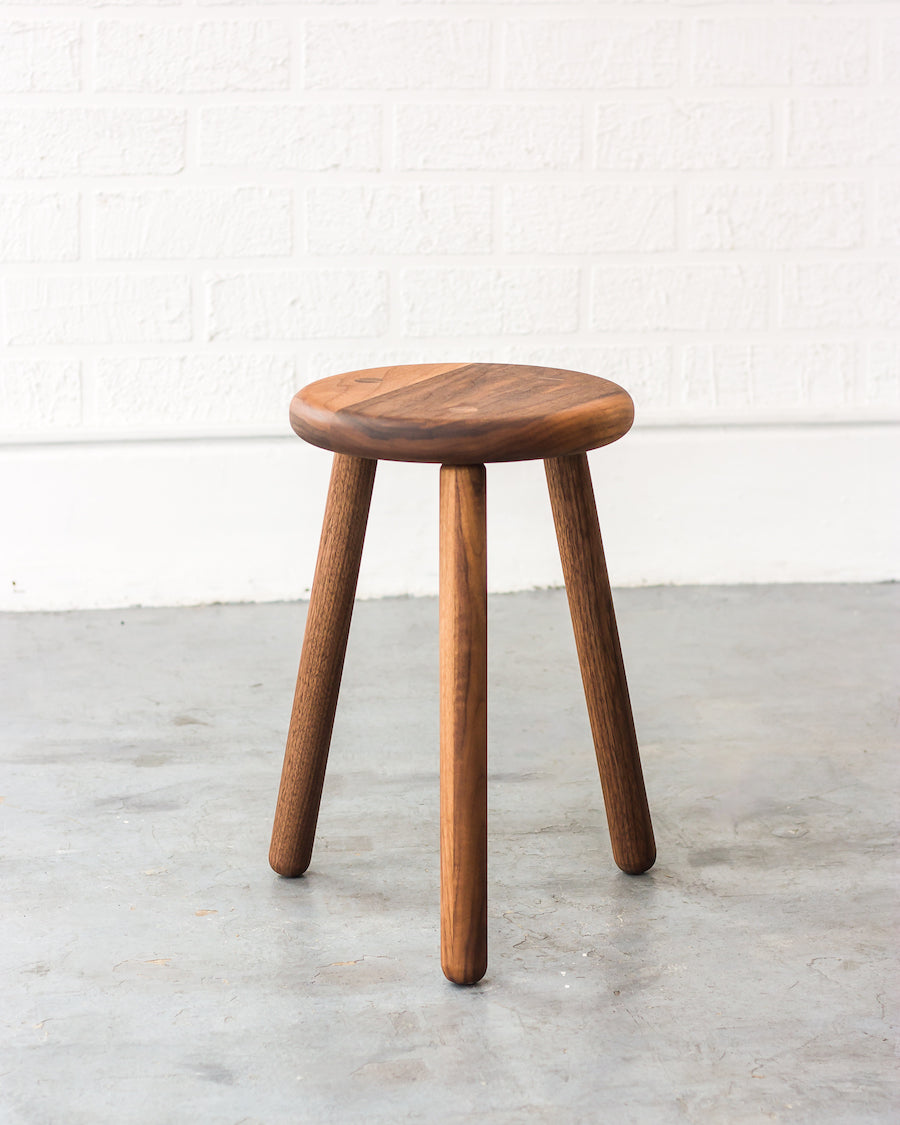 Wooden stool with three legs on a concrete floor against a white wall