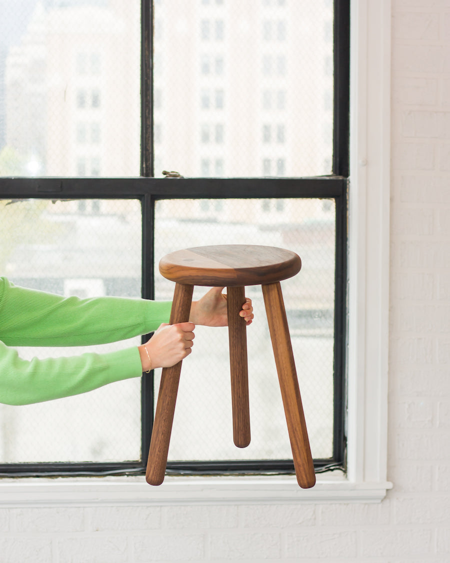 Person holding a wooden stool in front of a window with a cityscape view.