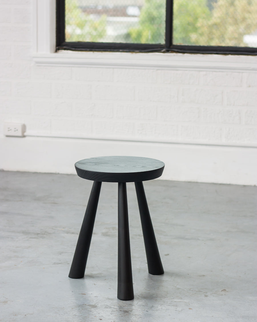 Black stool with a round seat in a room with white walls and a window.