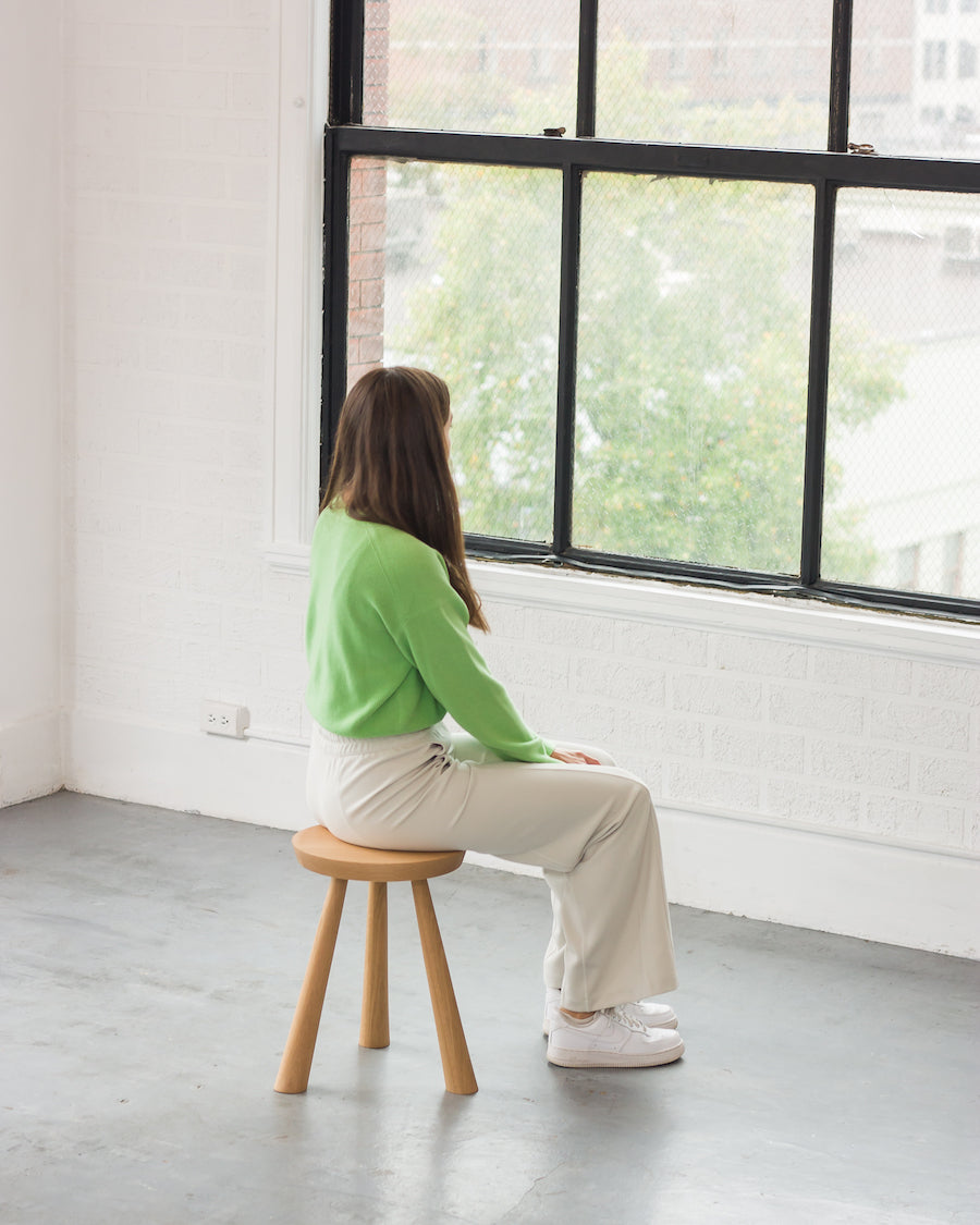 Person sitting on a wooden stool in a room with large windows.