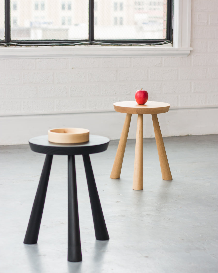 Two small round tables, one black and one wooden, with a bowl and an apple on a light gray floor.