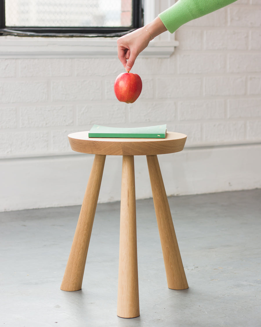 Person placing an apple on a small wooden stool with a white wall and window in the background