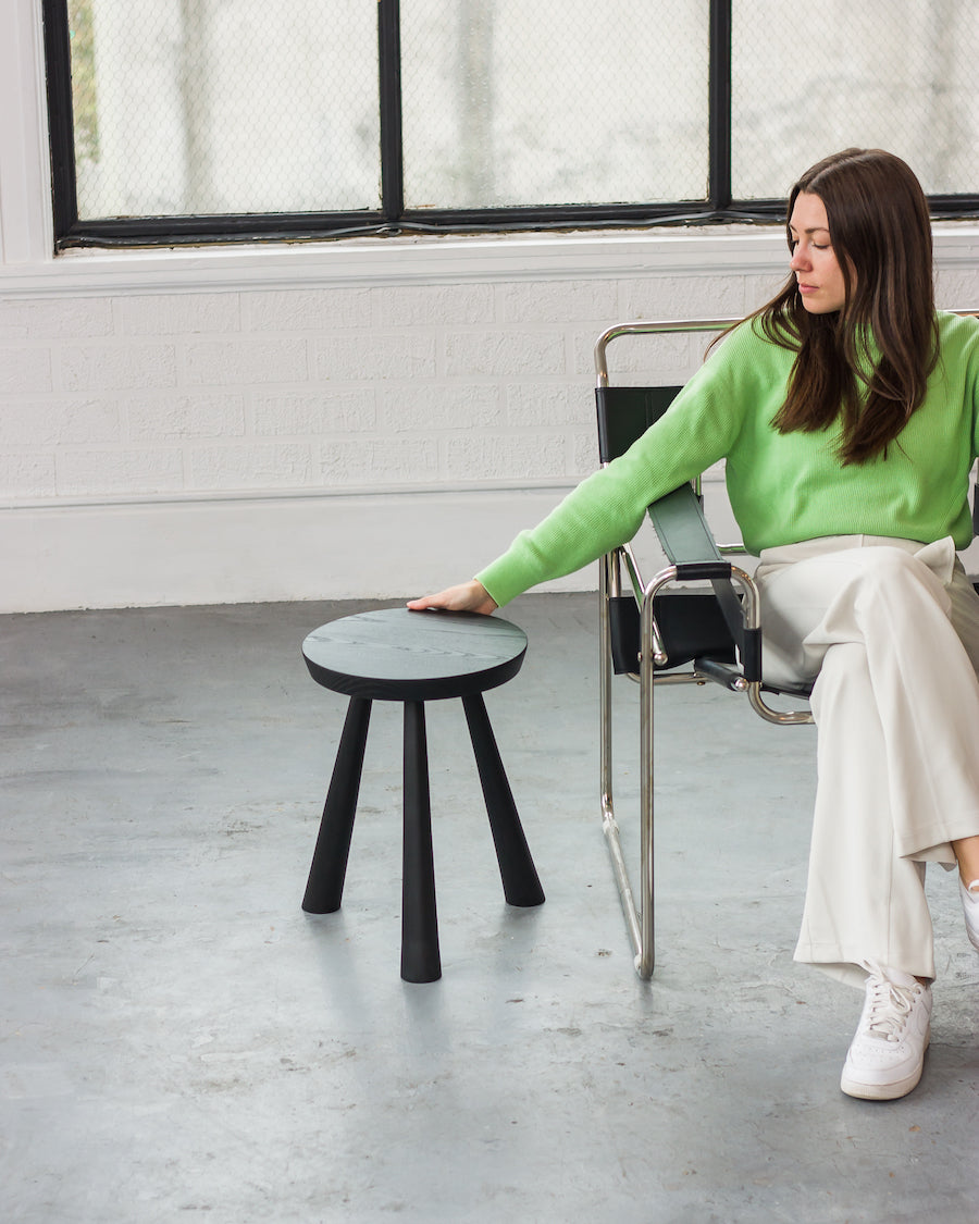 Woman in a green sweater sitting on a chair next to a black stool in a room with large windows.