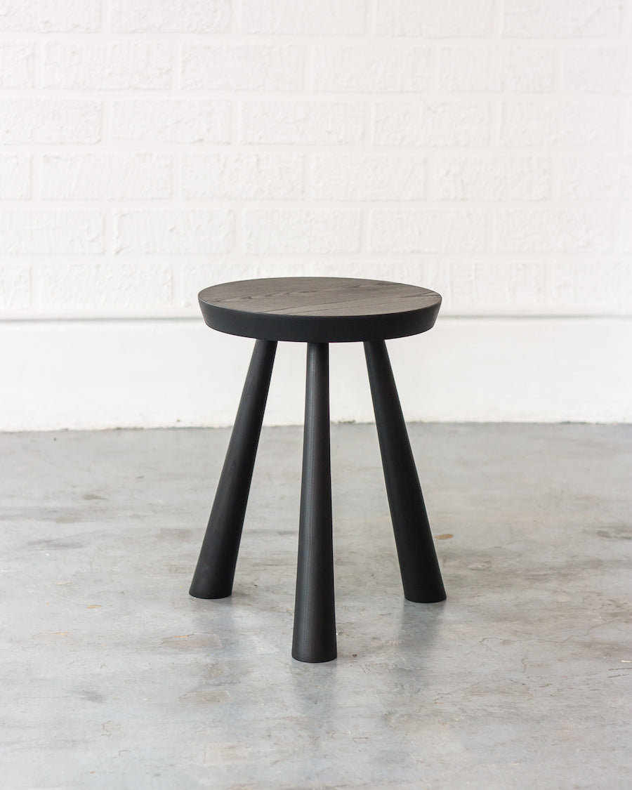 Black stool with a round seat on a light wooden floor against a white brick wall.