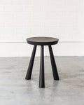 Black stool with a round seat on a light wooden floor against a white brick wall.