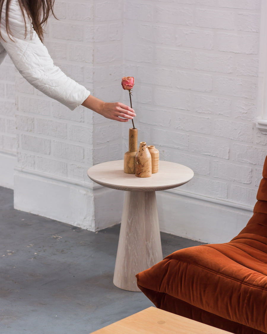 Person placing a rose in a wooden vase on a small round table with a white brick wall and brown chair in the background.