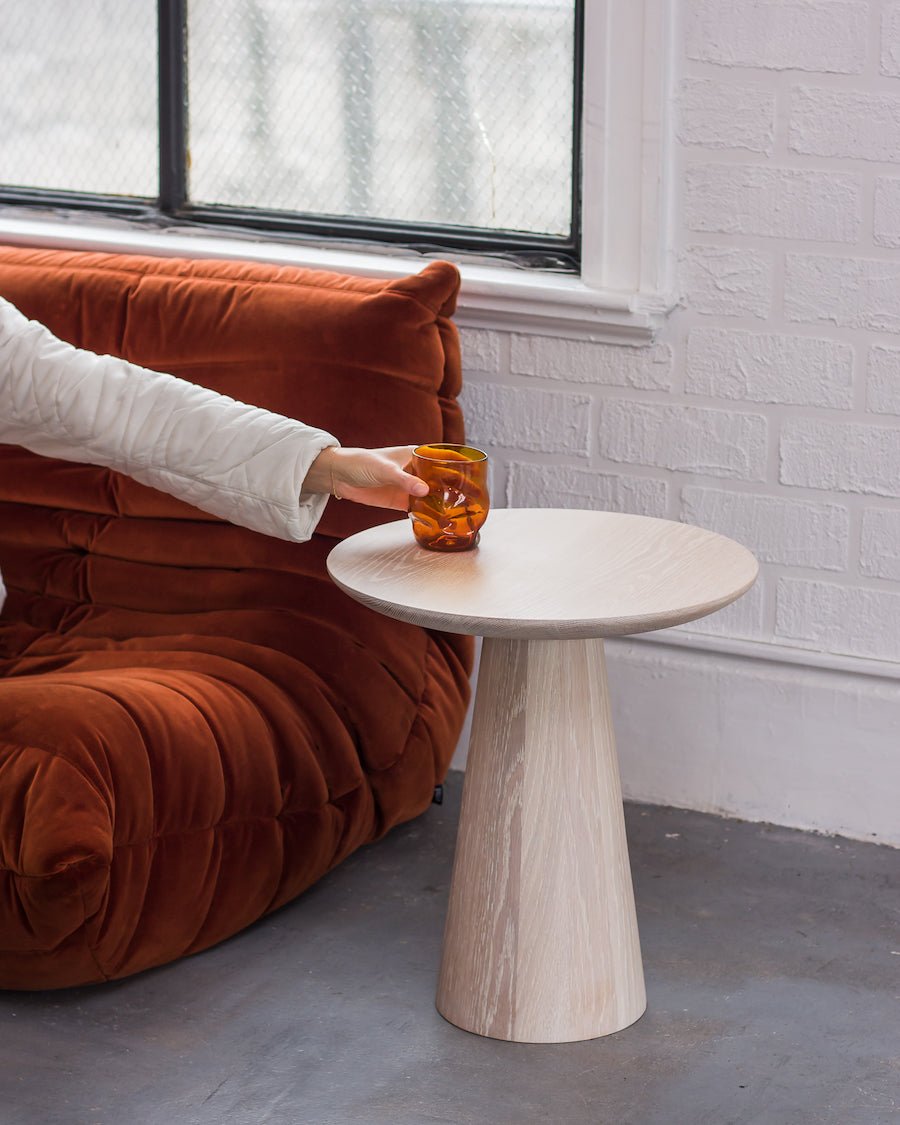 Person holding a glass on a small round wooden table next to a brown sofa.