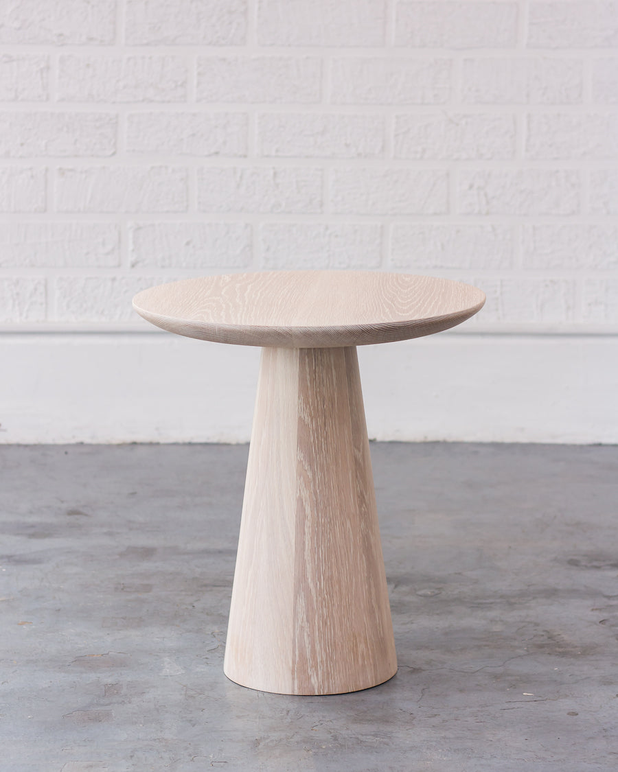 Wooden side table with a cylindrical base and round top against a white brick wall.