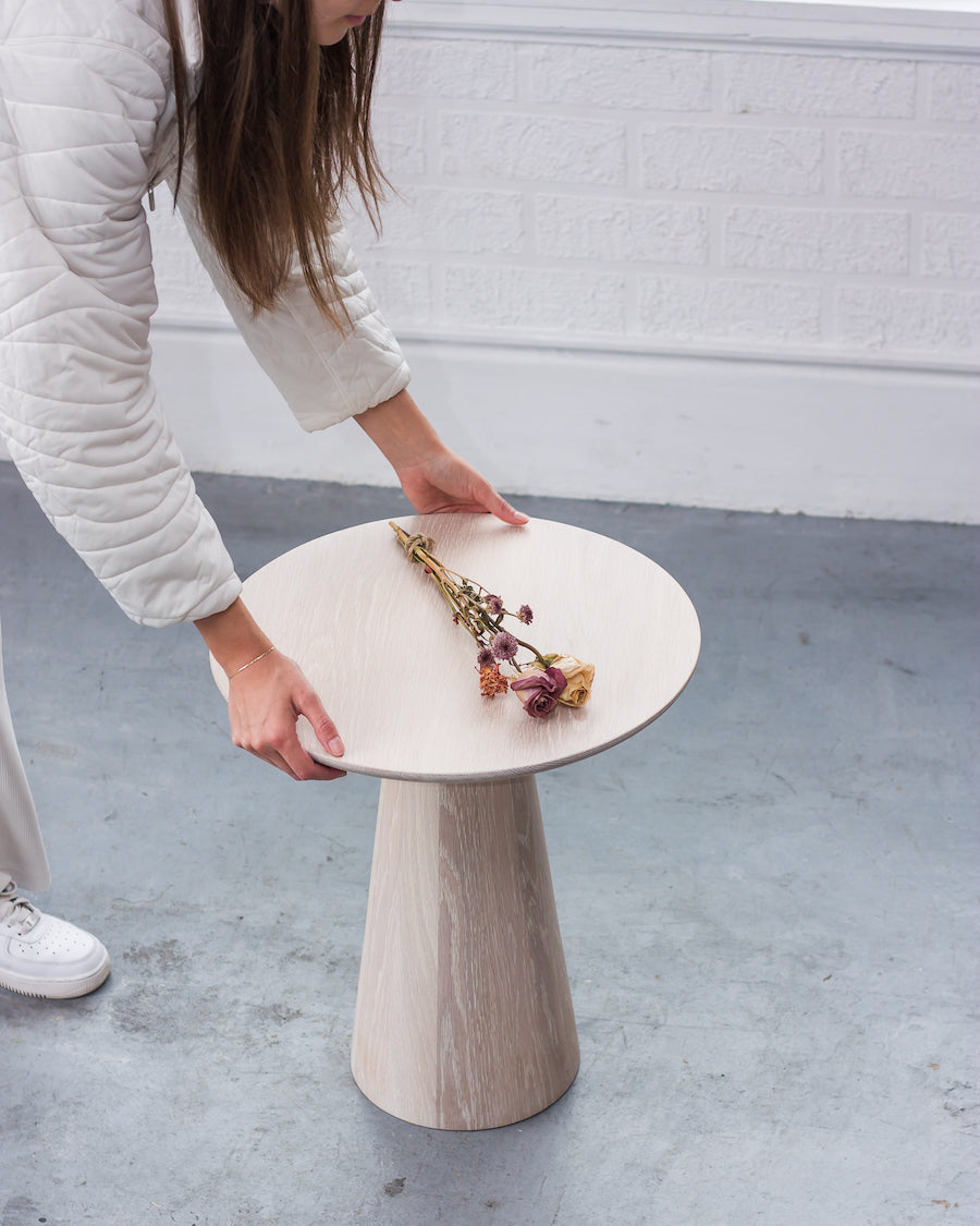 Person holding a small round table with a white top and wooden base, on a concrete floor.