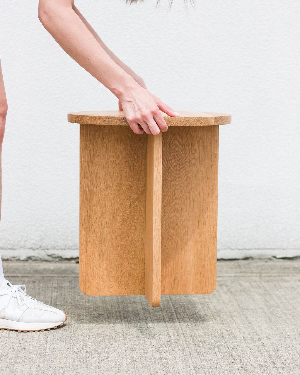 Wooden stool being held by a person on a textured floor with a white wall background