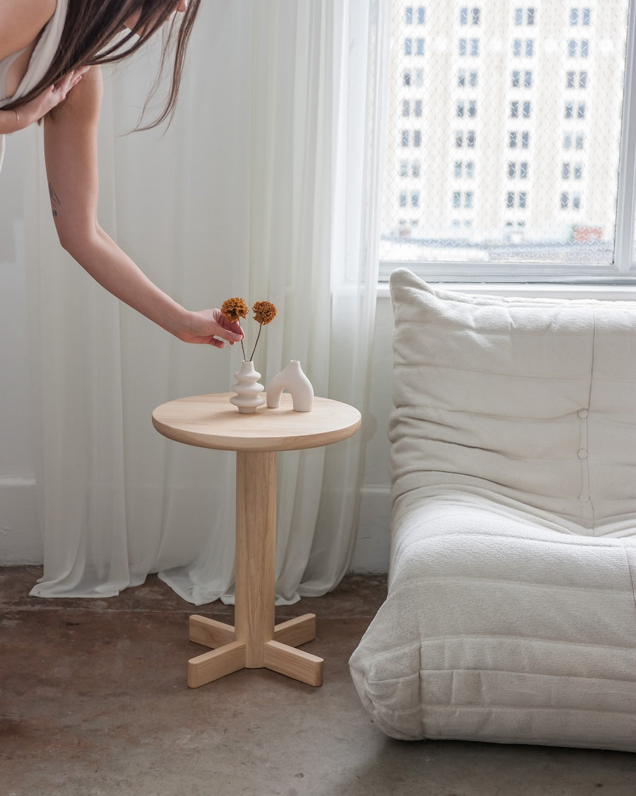 Person arranging flowers on a small wooden table in a room with a window and white curtains.