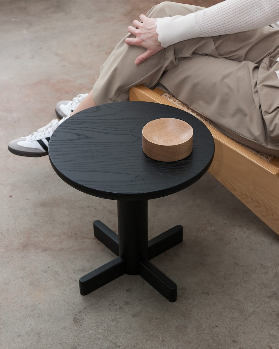 Black side table with a wooden bowl on a concrete floor, person sitting on a bed in the background.