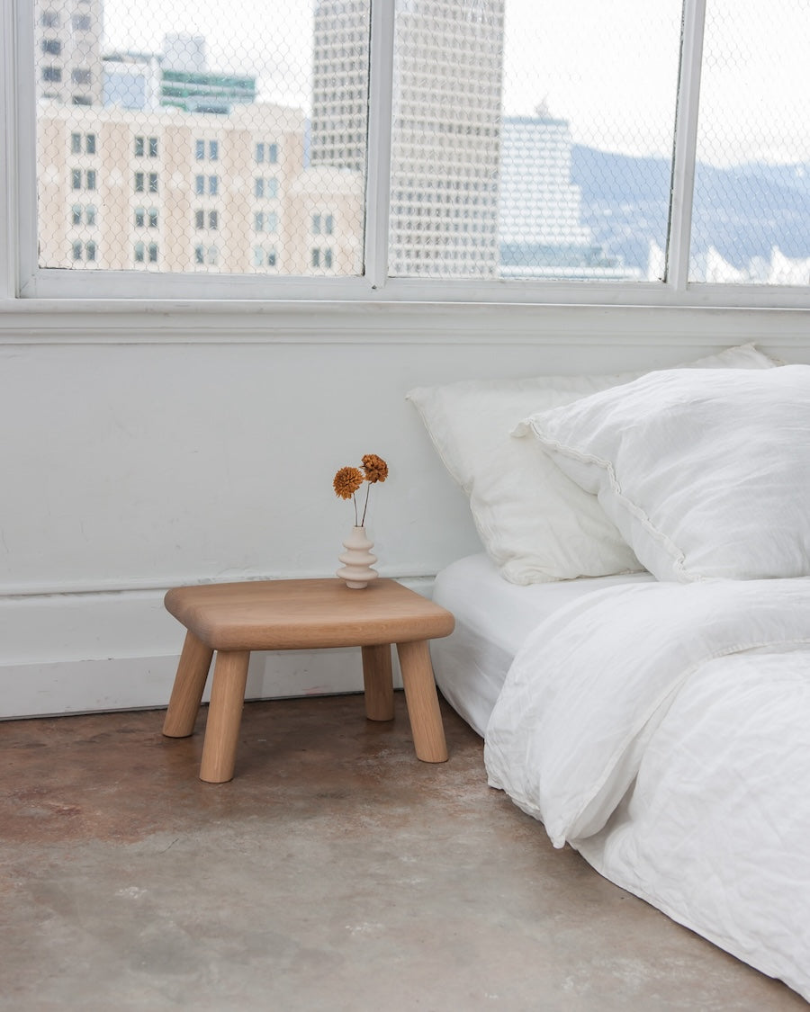 Wooden stool next to a bed with white bedding in a room with large windows.