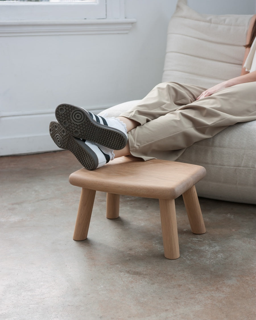 Person sitting on a wooden footstool with feet up, wearing black shoes and white socks, in a room with a beige sofa and concrete floor.