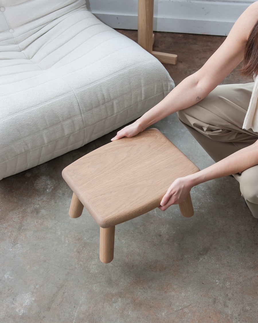 Person using a small wooden stool next to a sofa on a concrete floor.