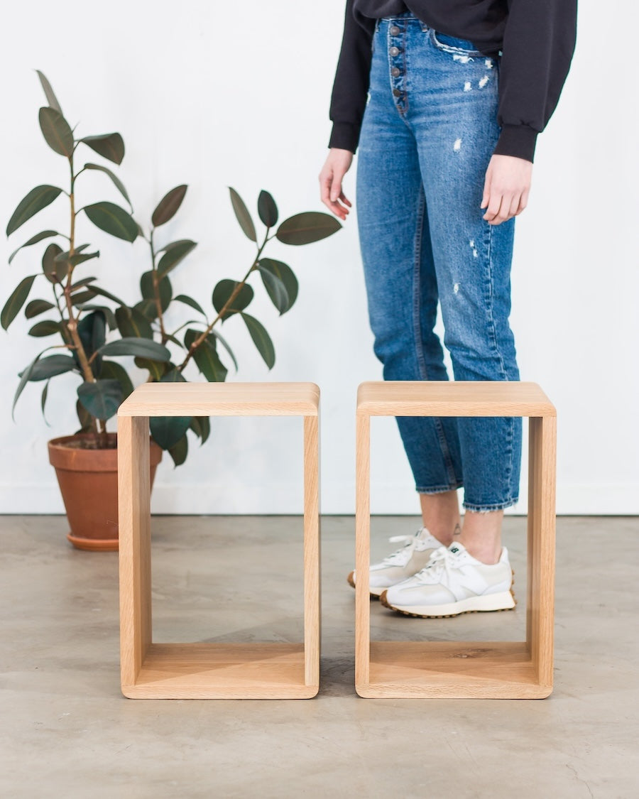 Two wooden stools on a light wood floor with a person standing behind them and a plant to the left.