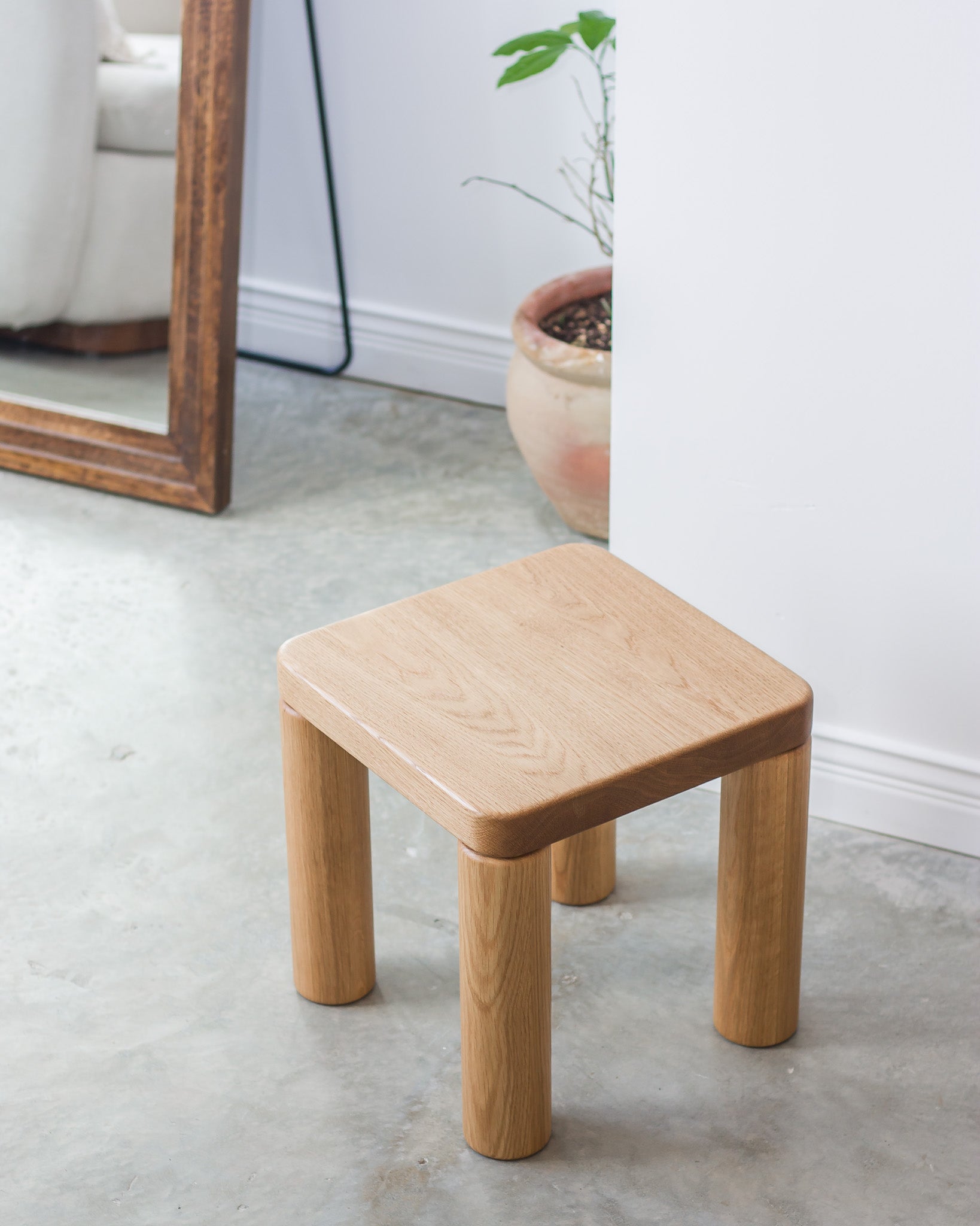Wooden stool on a concrete floor with a white wall and plant in the background