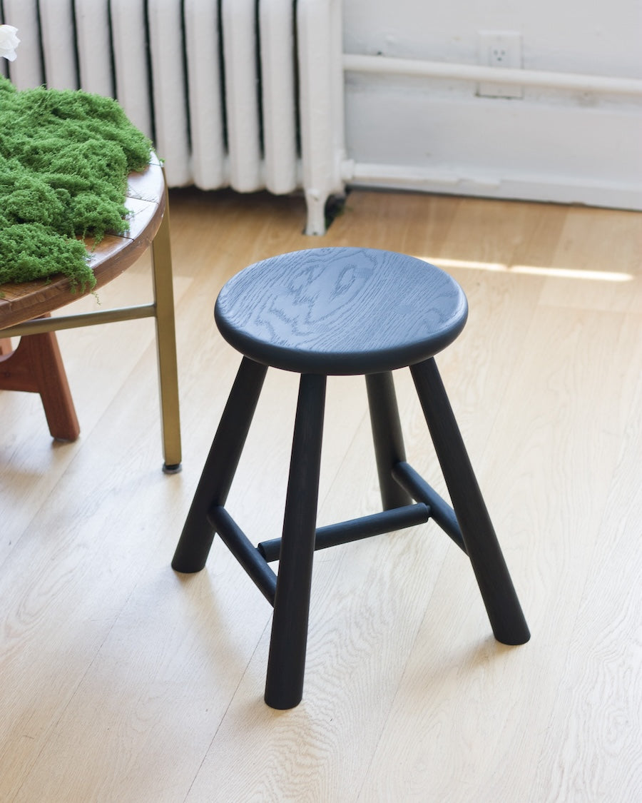 Black stool on a light wooden floor with a white wall and radiator in the background.