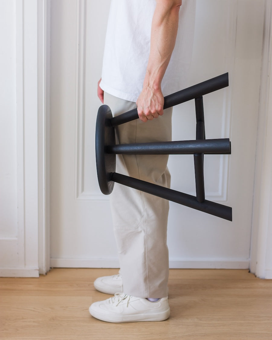 Person holding a black metal stool against a white wall.